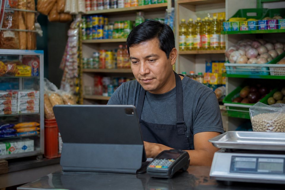Dueño latino de tienda de abastos gestionando ventas y cobranza en tableta
